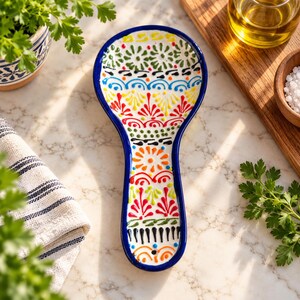 May include: A ceramic spoon rest with a white base and a dark blue rim, featuring a hand-painted pattern of colorful red, green, orange, and blue floral and geometric motifs arranged in horizontal bands along the elongated handle and bowl.