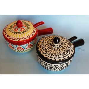 May include: Two ceramic bowls with lids and handles. The bowl on the left has a red base with white, yellow, green, and blue floral designs. The bowl on the right has a black base with white floral designs.
