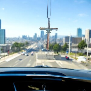 May include: A silver cross car charm hanging from the rearview mirror of a car. The cross is made of wire and has a beaded design. The cross is hanging in front of a blurred city background.