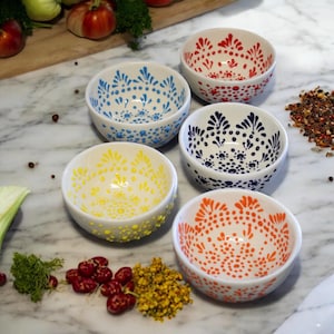 May include: Set of four small white ceramic bowls with colorful polka dot designs. The bowls are arranged in a circle on a marble surface. The colors of the bowls are blue, yellow, red, and orange.