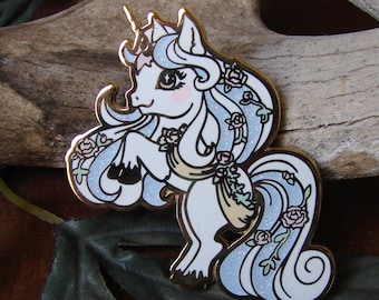 May include: A white unicorn pin with a gold outline and blue and pink floral accents. The unicorn has a flowing mane and tail with glitter accents.