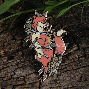Photo of an enamel pin of a fox with a bag of foraged mushrooms, a ribbon, sunflowers, and delphinium flowers