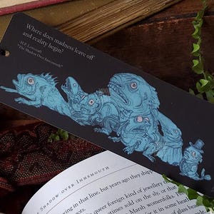 May include: A black rectangular bookmark featuring a blue illustration of multiple fish-like creatures and the text: Where does madness leave off and reality begin? H.P. Lovecraft The Shadow Over Innsmouth. A black cord is attached to the top left corner.