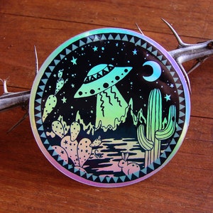 May include: A round sticker with a black background and a holographic finish. The sticker features a cartoon UFO flying over a desert landscape with cacti, a crescent moon, and stars. The sticker has a tribal-inspired border.