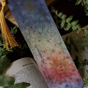 May include: A rectangular bookmark featuring a gradient design transitioning from deep blue at the top to warm orange at the bottom. The surface displays a celestial illustration with a sun, stars, and a tree, finished with a gold-colored tassel.