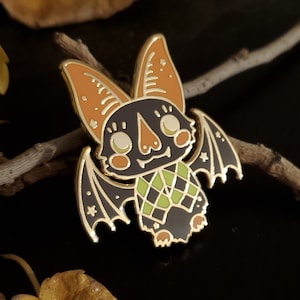 Puede incluir: Un pin de esmalte dorado con un murciélago de dibujos animados con orejas naranjas, alas negras y una bufanda con estampado verde y negro. El murciélago tiene una expresión amigable con ojos grandes y una pequeña boca.