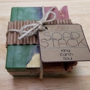May include: A stack of three handmade soap bars wrapped in brown paper and tied with twine. The soap bars are green, blue, and red. A brown paper tag is attached to the twine with the text "Soap Stack" and the words "King, Earth, Soul".