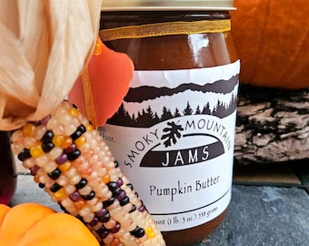 May include: A glass jar of pumpkin butter with a white label that reads "Smoky Mountain Jams Pumpkin Butter pint (1 lb. 3 oz.) 538 grams". The jar is sitting on a wooden surface with a small orange pumpkin and a colorful corn cob.