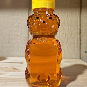 May include: A clear, bear-shaped honey bottle filled with golden honey. The bottle has a yellow cap and is made of transparent plastic. The bear's face is molded into the plastic, giving it a friendly appearance. The bottle is sitting on a wooden surface.