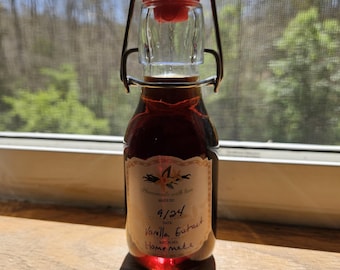 May include: A small glass bottle with a red and white stopper filled with a dark brown liquid. The bottle has a label that says "Homemade with love" and "9/24 Vanilla Extract Home Made".