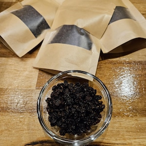 May include: A clear glass bowl filled with dark, dried berries sits on a wooden surface. Several small, brown paper pouches with a clear window are arranged around the bowl, also containing the same dark berries. The berries appear to be a deep, rich color.