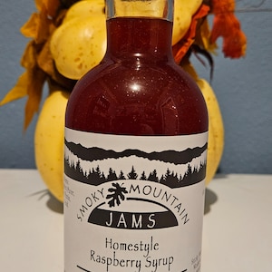 May include: A glass bottle of red raspberry syrup with a white label that reads "Smoky Mountain Jams Homestyle Raspberry Syrup 1 pint (1 lb. 4 oz.) 566 grams".