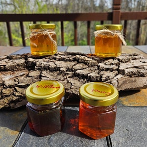 May include: Four small glass jars with gold lids filled with honey. The jars are labeled with red writing: "Wildflower", "Locust", and two unreadable labels. The jars are sitting on a piece of tree bark.