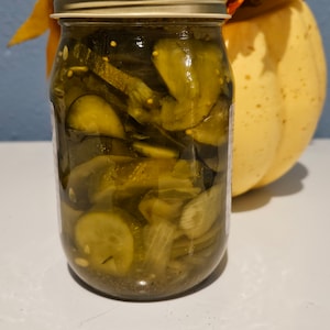 May include: A glass jar filled with green dill pickle slices. The jar has a silver lid and is sitting on a white surface.