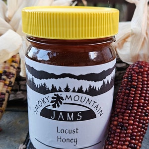 May include: A glass jar with a yellow lid filled with honey. The label reads "Smoky Mountain Jams Locust Honey 16 oz. (454 grams!)"