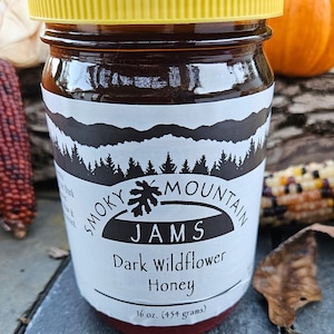 May include: A jar of Smoky Mountain Jams Dark Wildflower Honey with a yellow lid. The label features a mountain and forest design. The jar contains 16 oz (454 grams) of honey.