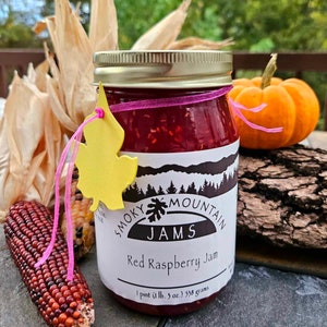 May include: A glass jar of red raspberry jam with a gold lid and a white label that reads "Smoky Mountain Jams Red Raspberry Jam 1 pint (1 lb. 3 oz.) 538 grams".