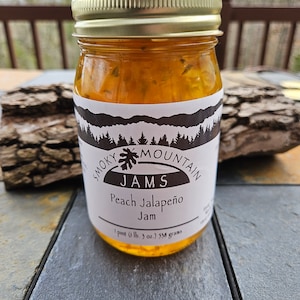 May include: A glass jar of Smoky Mountain Jams Peach Jalapeno Jam. The jar is labeled with a black and white logo and the text "Smoky Mountain Jams Peach Jalapeno Jam 1 pint (1 lb, 9 oz) 358 grams".
