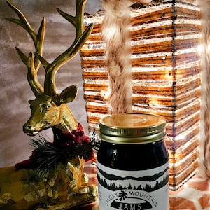 May include: A glass jar of Smoky Mountain Jams Black Raspberry Jam. The jar is sitting on a patterned surface with a gold deer figurine and a decorative light in the background.