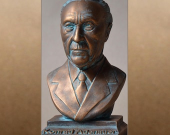 May include: A bronze bust of Konrad Adenauer, a German statesman, on a base with his name inscribed. The bust is a realistic depiction of Adenauer, with a weathered and aged appearance.