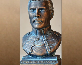 Könnte beinhalten: Eine Bronzestatue von Freddie Mercury, dem Leadsänger von Queen, mit einem detailliert modellierten Gesicht und einem strukturierten, geschichteten Oberkörper. Die Statue steht auf einem rechteckigen Sockel mit der Aufschrift "Freddie Mercury" auf der Vorderseite.