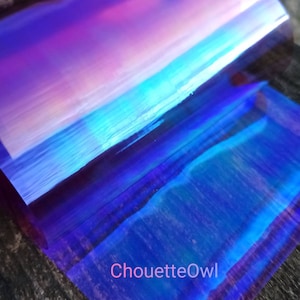 Puede incluir: Una lámina de vinilo holográfico iridiscente con un cambio de color azul, morado y rosa. La lámina está doblada por la mitad y el texto "ChouetteOwl" es visible en la esquina inferior derecha.