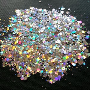Può includere: Primo piano di un mucchio di glitter iridescenti. I glitter sono composti da molti piccoli pezzi esagonali che riflettono un arcobaleno di colori. I glitter sono argentati e hanno un effetto olografico, con lampi di rosa, blu e verde.