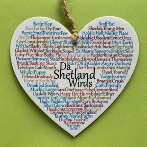 May include: A white heart-shaped wooden sign with the words "Da Shetland Wirds" in the centre. The sign is decorated with a variety of colourful words and phrases related to the Shetland Islands.