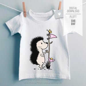 Könnte beinhalten: Weißes Kinder-T-Shirt mit einem Cartoon-Igel, der einen Holzstab mit einer rosa Fahne und einem gelben Schmetterling hält. Der Igel hat schwarze Stacheln und einen hellrosa Bauch. Das T-Shirt hängt an einer Wäscheleine mit einer Wäscheklammer.
