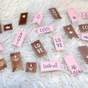 Könnte beinhalten: Eine Sammlung von braunen und rosafarbenen Stoff-Patches mit verschiedenen Designs und Texten, darunter "Unikat", "AHOI", "LOVE", "COOL" und "Locke der Boss".