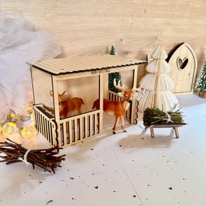 Könnte beinhalten: Ein hölzerner Rentierstall mit zwei Rentierfiguren, einem kleinen Heuwagen und dekorativen Bäumen. Der Stall trägt den Text "REINDEER STALL" auf dem Dach. Die Szene spielt auf einer weißen Oberfläche mit verstreuten schwarzen Herzformen.