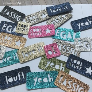 K&ouml;nnte beinhalten: Sortiment aus bunten Glitzeraufklebern mit verschiedenen W&ouml;rtern und S&auml;tzen, darunter "Cool Stuff", "Love It", "Yeah", "EGA", "Lassig" und "Classic".
