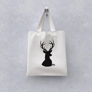 May include: A white tote bag with a black silhouette of a deer's head and antlers. The bag hangs from a metal hook against a light gray wall. The bag has a simple design, suitable for carrying groceries or personal items.