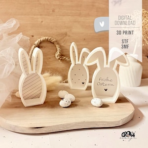 Puede incluir: Decoraciones de conejos de Pascua de madera color crema, con varios diseños, incluyendo rayas, lunares y el texto "Frohe Ostern". Los conejos están sobre una bandeja de madera con huevos moteados.