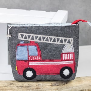 May include: A grey zippered pouch with a red fire engine applique. The engine has a white ladder, white stripes, and the word "TUTATA" embroidered on the side.