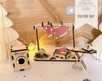 May include: A miniature wooden laundry set with a washing machine, clothesline, ironing board, and a small bed. The clothesline holds pink clothing. The image includes the text "DIGITAL DOWNLOAD LASER SVG PDF DXF".