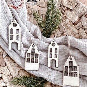 May include: Four wooden ornaments shaped like houses with cut-out windows and doors. The ornaments are hanging from twine and are on a light brown woven surface.
