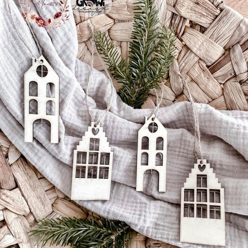 May include: Four wooden ornaments shaped like houses with cut-out windows and doors. The ornaments are hanging from twine and are on a light brown woven surface.