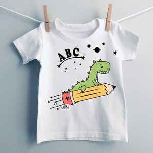 May include: White t-shirt with a green dinosaur riding a yellow pencil with a red eraser. The dinosaur is flying through space with stars and a planet. The text "ABC" is above the dinosaur.