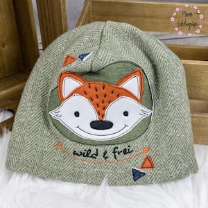 Op de afbeelding: Groene en bruine gebreide muts met een vos-applicatie en de tekst "wild & frei".