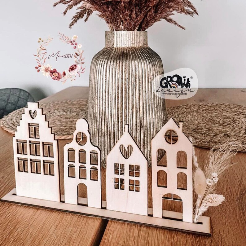 May include: A set of four wooden houses with cut-out windows and doors, arranged in a row on a wooden base. The houses are painted in a natural wood color and have a heart-shaped cutout on the front. The houses are decorated with dried pampas grass and flowers.