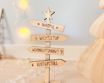 May include: A light-colored wooden signpost featuring a star at the top and four directional arrows. The arrows are engraved with the German text Nordpol, Wichteltür, Weihnachtsmarkt, and Rentierstall, along with small icons of stars, a door, and a candy cane.
