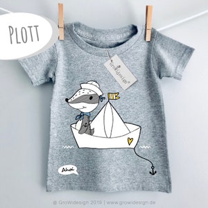 Könnte beinhalten: Ein graues T-Shirt mit einem Cartoon-Dachs mit Matrosenmütze und einem Seehund in einem Papierboot. Das Design enthält das Wort "Ahoi" in einer Sprechblase und das Wort "PLOTT" in einem weißen Oval. Ein Etikett mit dem Logo "Growidesign" ist angebracht.