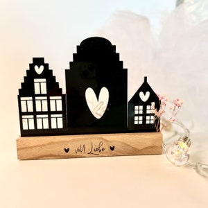 May include: Black silhouette of three houses with white hearts and windows on a wooden base with the text "voll Liebe" in black.