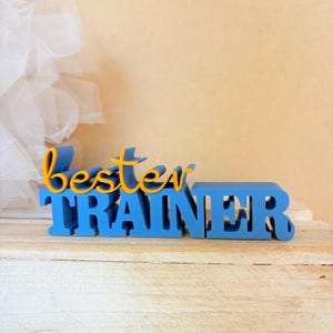 May include: A blue and yellow 3D-printed sign that reads "best trainer." The word "best" is in yellow script, and "TRAINER" is in bold blue block letters. The sign is on a wooden surface.
