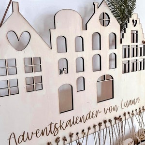 May include: Wooden advent calendar shaped like a row of houses with numbered bags hanging from the bottom. The calendar is personalized with the text "Adventskalender von Luana".