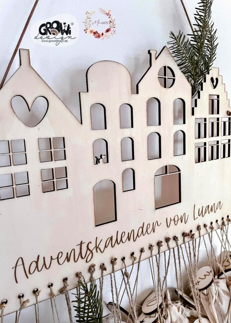 May include: Wooden advent calendar shaped like a row of houses with numbered bags hanging from the bottom. The calendar is personalized with the text "Adventskalender von Luana".