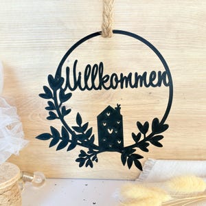 Könnte beinhalten: Schwarzer runder Wandschmuck mit dem Wort "Willkommen" und einer Haussilhouette. Das Design ist von einem Blätterkranz umgeben und wird von einem Juteseil gehalten. Der Hintergrund ist eine helle Holzoberfläche.