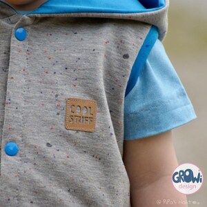 K&ouml;nnte beinhalten: Eine graue Kapuzenweste mit blauem Futter und einem braunen Patch mit der Aufschrift "COOL STUFF". Die Weste hat ein gesprenkeltes Muster und blaue Kn&ouml;pfe. Die Weste wird von einem Kind getragen.