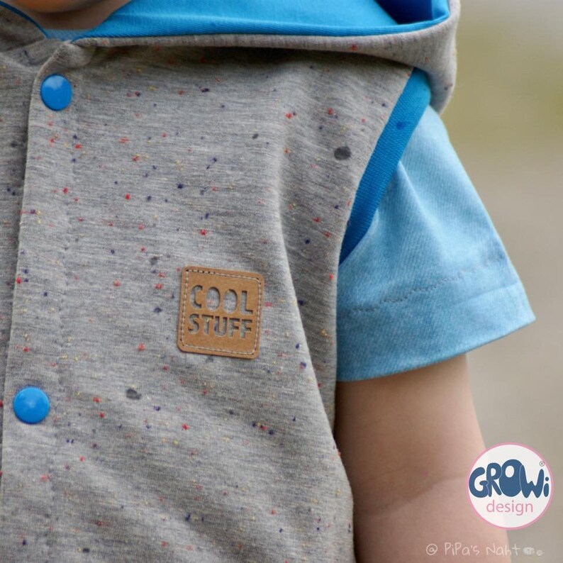 K&ouml;nnte beinhalten: Eine graue Kapuzenweste mit blauem Futter und einem braunen Patch mit der Aufschrift "COOL STUFF". Die Weste hat ein gesprenkeltes Muster und blaue Kn&ouml;pfe. Die Weste wird von einem Kind getragen.